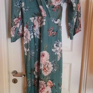 Grön blommig omlottklänning från NLY  - Säljer en lång grön omlottklänning från Only med vackert blommigt mönster i rosa och vitt. Klänningen har långa ärmar och markerad midja. Perfekt för vår och sommar, tillverkad i mjuk polyester.. Lång slits. Använd 2 gånger. Men sömmarna är synliga i midjan enda från början
