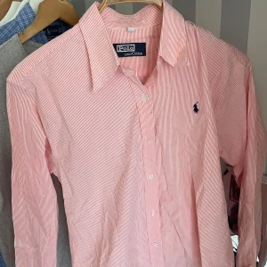 Orangevit randig skjorta från Polo - Klassisk skjorta från Polo i orangevitt randigt mönster. Skjortan är tillverkad i 100% bomull och har långa ärmar, knappar framtill och en broderad logga på bröstet. Perfekt för en stilren och tidlös look. Storleken som står i tröjan är xxl men den är M