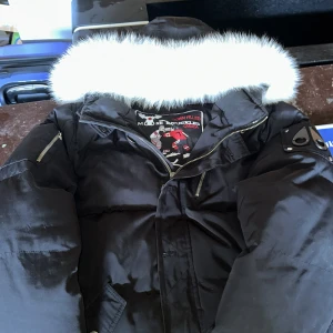 Svart dunjacka med pälskant från Moose Knuckles - Säljer en svart dunjacka från Moose Knuckles med vit pälskant på huvan. Jackan har dragkedja, flera fickor och en snygg logga på ärmen. Perfekt för kalla dagar och riktigt varm.