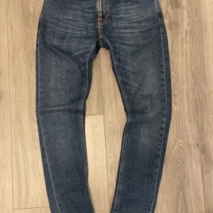 Blå jeans från Nudie Jeans - Säljer ett par klassiska blå jeans från Nudie Jeans med orangea sömmar och femficksmodell. Byxorna har normal passform och raka ben. Perfekta för dig som gillar en tidlös stil.