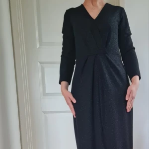 Svart omlottklänning med v-ringning - Elegant svart klänning med omlott-design och v-ringad hals. Klänningen har långa ärmar och en figurnära passform upptill med lätt drapering framtill. Perfekt för dig som gillar stilrena och klassiska plagg.Tyget är glittrigt om det inte framstår på bilden..Om det behövs kan jag skicka bättre bilder på klänningen vid intresse 