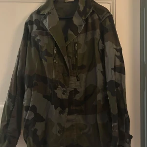 Camouflagejacka med dragkedjor från Quatre filles - Säljer en grön och svart camouflage vindjacka med krage och knäppning framtill. Jackan har långa ärmar och två dragkedjefickor på bröstet. 