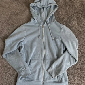 Ljusblå zip up hoodie - Säljer en enkel och stilren ljusblå hoodie med dragkedja och huva. Tröjan har långa ärmar, fickor framtill och justerbara snören i huvan. Perfekt för en avslappnad look.