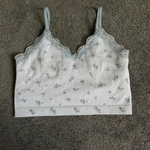 Vit blommig linnetopp med spets från Primark - Söt vit linnetopp från Primark med ljusblå småblommigt mönster och spetskant längs v-ringningen. Smala axelband och ribbad struktur ger en feminin look. Perfekt för varma dagar eller som lager under andra plagg.