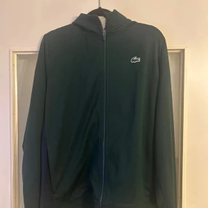 Mörkgrön hoodiejacka från Lacoste Sport - Mörkgrön hoodiejacka från Lacoste Sport med dragkedja framtill och klassisk krokodillogga på bröstet. Jackan har huva och är långärmad, perfekt för en sportig och avslappnad stil.