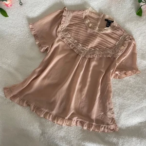 Ljusrosa blus med spetsdetaljer från forever 21 - Supersöt ljusrosa blus från forever 21 med volangkant och spetsdetaljer vid halsringning och ärmslut. Blusen har korta ärmar, knappar i ryggen och ett romantiskt, luftigt fall. Perfekt för vår och sommar!