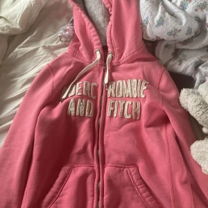 Rosa hoodie från Abercrombie & Fitch - Säljer en rosa hoodie med dragkedja och huva från Abercrombie & Fitch. Tröjan har vita snören och broderad logga framtill. Perfekt för en avslappnad stil.
