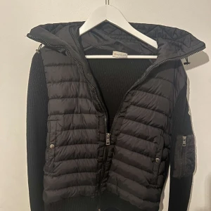 Moncler Cardigan - Säljer en cardigan från Moncler med stickade ärmar och huva. Jackan har dragkedja framtill och en ficka med dragkedja på ärmen. Klassisk Moncler-logga på vänster ärm. Perfekt för vår, höst, varmare vinter och sommarkvällar. M passar S