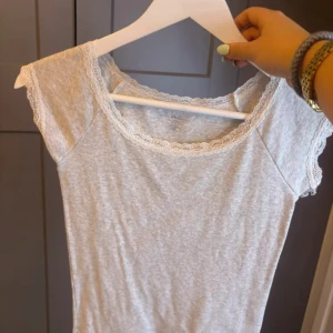 Ljusgrå topp med spetsdetaljer - Säljer en ljusgrå topp med korta ärmar och fina spetsdetaljer längs halsringningen och ärmsluten. Toppen är i mjukt bomullsmaterial och har en feminin och enkel design som passar perfekt till vardags.