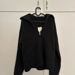 Mörkgrå stickad tröja med dragkedja - Mysig mörkgrå stickad tröja från Bikbok med halv dragkedja framtill och krage. Tröjan har lång ärm och är tillverkad av återvunnen polyester. Perfekt för kyliga dagar och enkel att matcha med olika outfits.