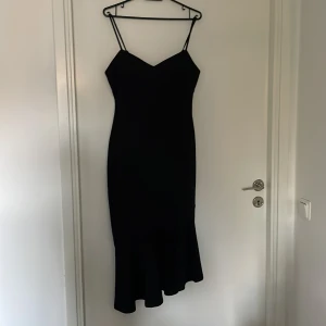 Svart klänning med volang från Bubbleroom - Supersnygg svart klänning från Bubbleroom med smala axelband och volang nertill. Modellen är figurnära och har en elegant v-ringad urringning. Helt oanvänd och har storleken S.
