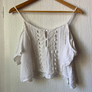 Vit croppad blus med spetsdetaljer - Säljer en vit croppad blus med tunna axelband och offshoulder-ärmar. Framsidan har dekorativa spetsdetaljer och knappar. Perfekt för varma dagar och somriga outfits.