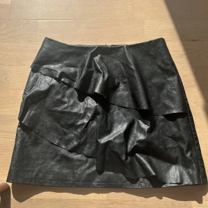 Svart kjol med volang från Lindex - Säljer en svart  kjol från Lindex med snygg volangdetalj framtill. Kjolen har en rak passform och är tillverkad i ett material som liknar skinn. Perfekt för dig som vill ha en trendig och stilren look.