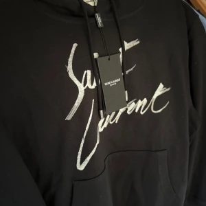 Svart hoodie från Saint Laurent - Säljer en svart hoodie från Saint Laurent med vit logotyp framtill. Tröjan har huva med dragsko och en stor magficka. Perfekt för dig som gillar stilrena och exklusiva plagg.