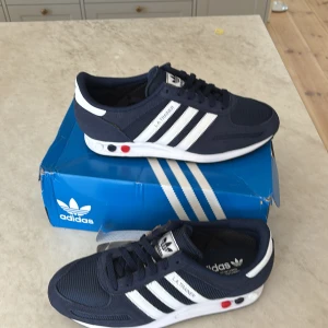 Adidas LA Trainer sneakers i marinblått - Säljer ett par Adidas LA Trainer sneakers i marinblått med vita ränder och detaljer. Skorna har snörning, platt sula och klassisk retrostil. Perfekta för dig som gillar sportig och stilren look. Levereras med originalkartong.