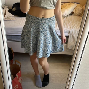 Blå blommig kortkjol från 157 - 20kr Söt blå kortkjol från 157 med vitt och svart blommönster. Kjolen har resår i midjan och är lätt utsvängd för en skön passform. Perfekt för varma dagar och enkel att matcha med olika toppar.