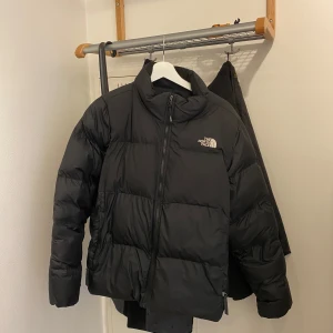 Svart dunjacka från The North Face - Säljer en svart dunjacka från The North Face med hög krage och dragkedja framtill. Jackan är vadderad och har en klassisk pufferstil med logga på bröstet. Perfekt för kyliga dagar och enkel att matcha med olika outfits.