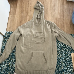 Beige stickad hoodie med S-brodyr - Säljer en beige stickad hoodie med stor broderad bokstav S på bröstet. Tröjan har huva, lång ärm och en klassisk känguruficka framtill. Den har aldrig kommit till användning så säljer den