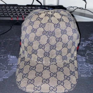 Beige keps från Gucci med monogram - Säljer en beige keps från Gucci med klassiskt GG-monogram i mörkblått och röd-blå rand på sidan. Kepsen har böjd skärm och justerbar passform. Perfekt för dig som gillar exklusiva accessoarer. Haft den ett hyfsat bra tag och använder inte den längre. Jag är även på för byte  