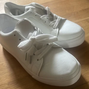 Vita sneakers med snörning - Säljer ett par klassiska vita sneakers med rund tå och platt sula. Skorna har breda vita skosnören och är tillverkade i syntetmaterial. Perfekta för en clean och stilren look.