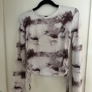 Långärmad croppad topp med abstrakt mönster - Säljer en långärmad croppad topp med dragsko i sidorna. Toppen har ett abstrakt mönster i brunt och vitt, och är tillverkad i ett mjukt, stretchigt material. 