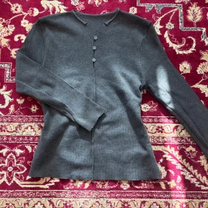 Grå stickad kofta med knappar - Säljer en stilren grå stickad kofta med tre knappar framtill och lång ärm. Koftan har en enkel och klassisk design med lätt v-ringad halsringning. Perfekt att slänga på sig när det blir lite kyligt.