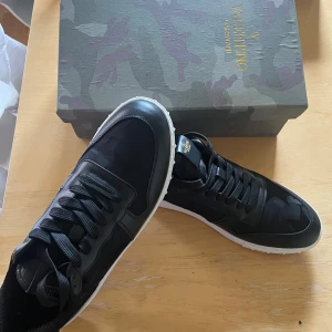 Svarta Valentino Garavani sneakers - Säljer ett par svarta Valentino Garavani sneakers med vita sulor och diskret kamouflagemönster. Skorna har snörning och rund tå, samt detaljer i både skinn och syntet. Perfekta för dig som vill ha en stilren och exklusiv look. Dustbag och box medföljer!