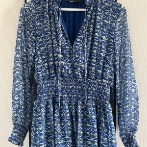 Blåmönstrad långärmad klänning från Zara - Säljer en blå och vit mönstrad klänning från Zara med smockad midja och långa ärmar. Klänningen har v-ringning och ett lätt, luftigt tyg som ger en fin siluett. Perfekt för vår och sommar!