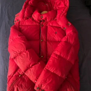 Röd dunjacka från Polo Ralph Lauren - Säljer en röd dunjacka från Polo Ralph Lauren med huva och dragkedja. Jackan har knappar, två stora fickor framtill och klassisk Polo-logga på bröstet. Perfekt för vår och sköna sommar dagar och riktigt snygg färg. 