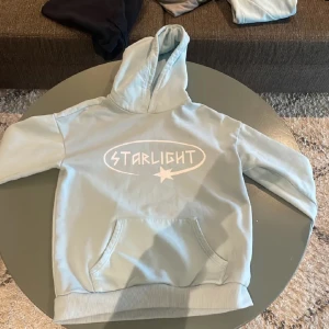 Ljusblå hoodie från Starlight - Säljer en ljusblå hoodie från Starlight med vit logga och stjärndetalj på bröstet. Tröjan har huva och en stor magficka framtill. Perfekt för en avslappnad och cool stil. Finns bara 50 i värden