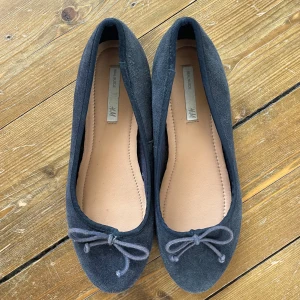 Mörkblå mocka ballerinaskor från H&M - Säljer ett par klassiska mörkblå ballerinaskor från H&M i äkta mocka. Skorna har rund tå och en liten rosett framtill för en söt touch. Perfekta att matcha med både jeans och klänning.