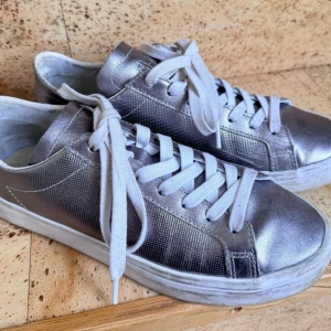 Silvriga sneakers från Adidas - Säljer ett par silvriga sneakers från Adidas med vit sula och vita skosnören. Skorna har en metallic finish och klassisk modell. Perfekta för dig som vill sticka ut med en glansig look. Lite högre sula. 