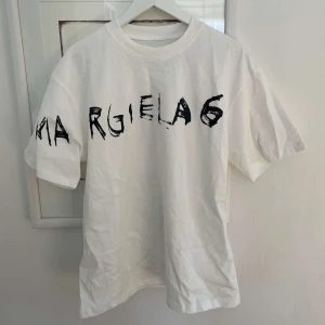 Vit t-shirt från Margiela 6 med tryck - Nu säljer vi en vit t-shirt från maison margiela med svart grafiskt tryck över bröstet och axeln. Perfekt för dig som gillar streetwear och unika designer.