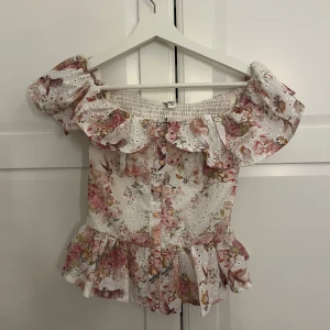 Blommig offshoulder korsett topp med volang - Söt blommig offshoulder korsett aktig topp med volangkant och knappar framtill. Blusen har broderade detaljer och är i vitt med rosa och gröna blommor. Smockad rygg och peplum nedtill ger en feminin siluett.