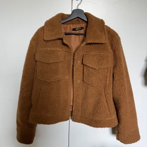Brun teddyjacka från Gina Tricot - Mysig brun teddyjacka från Gina Tricot med krage och två stora bröstfickor. Jackan är croppad och har knappar framtill. Perfekt för kyligare dagar och ger en trendig look. Storlek 38 men passar xs-m, endast testad💗