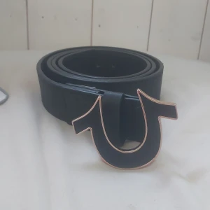 Svart bälte från True Religion med stor logga - Säljer ett svart bälte från True Religion med en stor, unik logga som spänne. Bältet är gjort i skinn och har tydlig märkning på insidan. Perfekt för dig som vill ha en statement-accessoar.