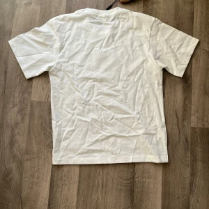 Vit t-shirt från Burberry med tryck - Säljer en vit t-shirt från Burberry med svart och guld texttryck på bröstet där det står 'BURBERRY LONDON ENGLAND'. Klassisk passform och rund halsringning. Perfekt för dig som gillar stilrena märkesplagg.