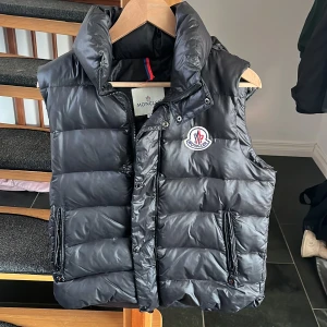 Moncler väst - Säljer en svart dunväst från Moncler med hög krage och två dragkedjefickor framtill. Västen har Moncler-logga på bröstet och klassisk quiltad design. Perfekt för lager-på-lager och stilren look. 