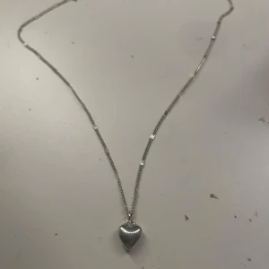 Silverfärgat halsband med hjärthänge - Ett stilrent halsband i silverfärg med ett litet hjärta som hänge. Kedjan är tunn och diskret, perfekt för att ge en söt detalj till din outfit. Passar dig som gillar enkla och klassiska smycken.