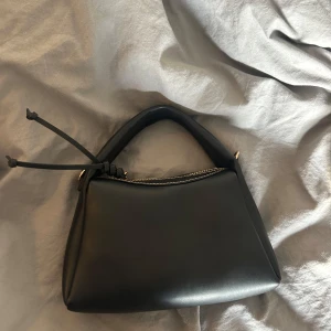 Svart handväska med dragkedja - Säljer en stilren svart handväska i slätt material med guldfärgade detaljer och dragkedja upptill. Väskan har en bred axelrem och dekorativa sömmar längs kanterna. Perfekt för att bära det viktigaste. Ifrån hm 