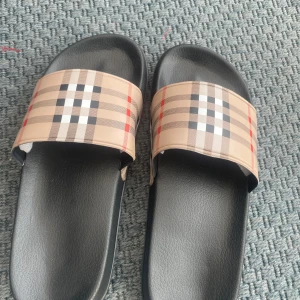 Burberry sandaler - Säljer ett par svarta sandaler med breda remmar i klassiskt beige-rutigt mönster. Perfekta för sommaren och riktigt sköna att glida runt i. Snygg design som sticker ut och passar till många outfits.