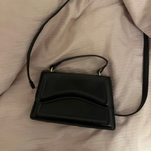 Svart axelväska  - Säljer en stilren svart axelväska med både axelrem och handtag. Väskan har en elegant, enkel design med mjuka former och guldiga detaljer vid remmarna. Den är väldigt rymlig, perfekt för att bära det viktigaste och passar till många olika outfits. 