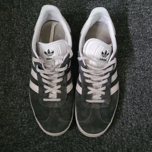 Adidas Gazelle sneakers i svart mocka - Säljer ett par klassiska Adidas Gazelle sneakers i svart mocka med vita ränder och vit sula. Skorna har snörning och den ikoniska Gazelle-loggan i guld på sidan. Perfekta för dig som gillar retrostil och streetwear.