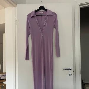 Lila långklänning från NA-KD - Lång, lila klänning från NA-KD med mjukt, stretchigt material. Klänningen har lång ärm, djup v-ringning med knappar framtill och en bred krage som ger en cool retrovibe. Perfekt för dig som vill sticka ut med en stilren look.