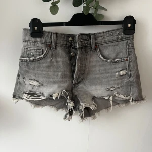 Grå slitna jeansshorts från Zara - Snygga grå jeansshorts från Zara med medel/låg midja, fransiga kanter. Shortsen har slitningar och trasiga detaljer för en cool, edgy look. Klassisk femficksmodell med knappgylf och en tvättad grå färg som passar perfekt till sommaren.
