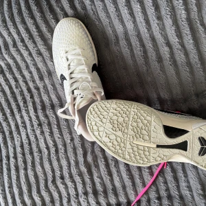 Vita Nike Kobe basketskor med stjärn mönster - Säljer ett par vita Nike basketskor med coolt strukturmönster på ovandelen och svart swoosh. Ena skon har vita snören och den andra har neonrosa snören för extra attityd. Skorna har platt sula och är perfekta för inomhusspel.