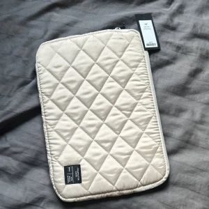 Quiltad beige datorväska 13 tum - Snygg quiltad datorväska från Arket i beige för 13-tums laptop. Mjuk och vadderad med dragkedja runt kanten. Perfekt för att skydda din dator och ge en clean look. Minimalistisk design med diskret svart etikett i hörnet. Aldrig använd, prislappen sitter kvar.