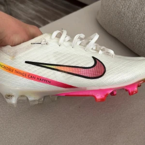 Nike Mercurial vita fotbollsskor - Nike Mercurial fotbollsskor i vitt med färgglada detaljer i rosa, orange och gult. Swoosh-logga i svart och rosa, texten 'Incredible things can happen' på sidan. Ovandel i syntetmaterial med snörning och platt sula med dobbar i rosa och orange. Perfekta för dig som vill sticka ut på planen.
