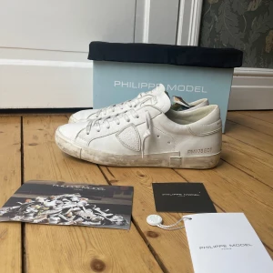 Vita Philippe Model sneakers  - Snygga vita sneakers från Philippe Model i äkta skinn med klassisk låg profil och rund tå. Skorna har diskret logga på sidan och häl, samt vita snören. Perfekta för dig som gillar stilrena och trendiga sneakers med exklusiv känsla.
