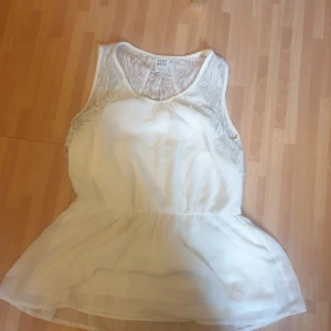 Vit spetsblus från Vero Moda - Supersöt vit blus från Vero Moda med spetsdetaljer upptill och på ryggen. Blusen är ärmlös och har en lätt utsvängd nederdel som ger en flowy känsla. Perfekt för dig som gillar romantisk och feminin stil.